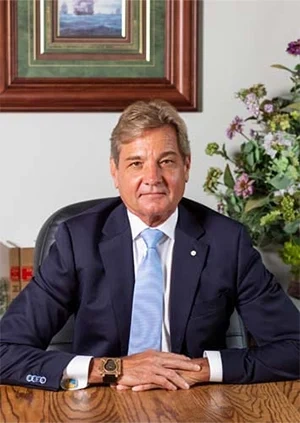 Man in a navy suit with a light blue tie sits at a wooden desk in an office, hands folded, with framed art and flowers in the background.