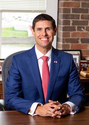 Professional portrait of a man in a blue suit and red tie seated at a desk in an office, hands folded.